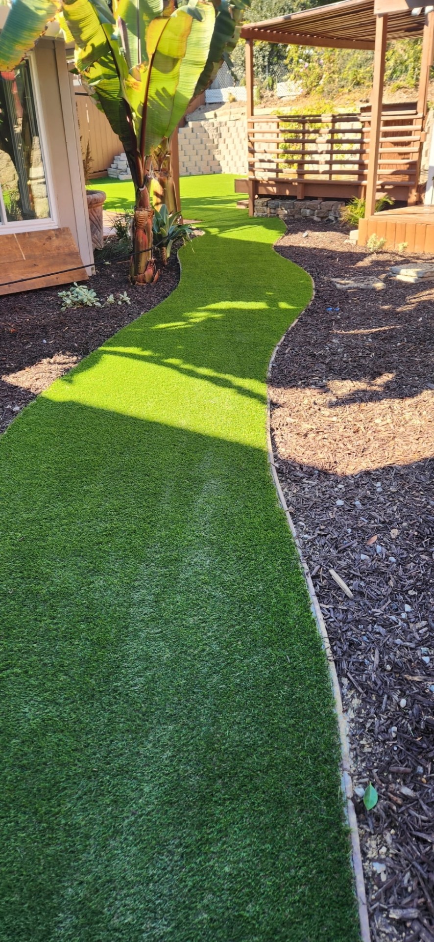 Curved artificial turf pathway through a backyard with pergola and mulch beds